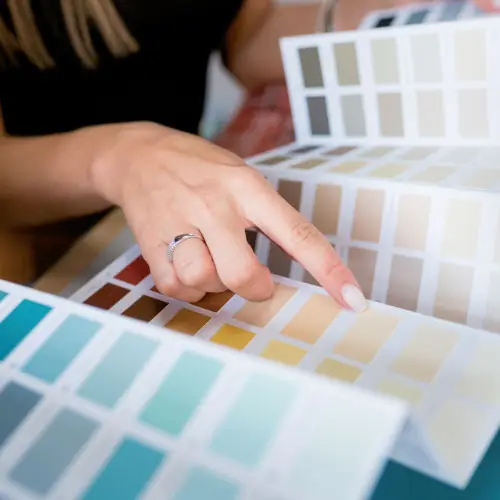 A hand pointing to a light beige shade on a detailed color swatch book, which contains an array of paint colors, including various shades of blue, teal, beige, and brown. The person is wearing a silver ring.
