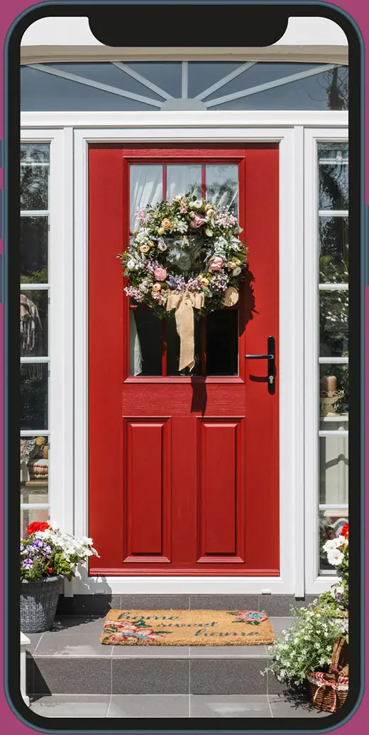 A vibrant red front door with decorative panels, adorned with a lush floral wreath featuring roses, pastel blooms, and greenery, accented by a beige ribbon. The door is framed by white trim and flanked by side windows, with potted flowers on the doorstep.